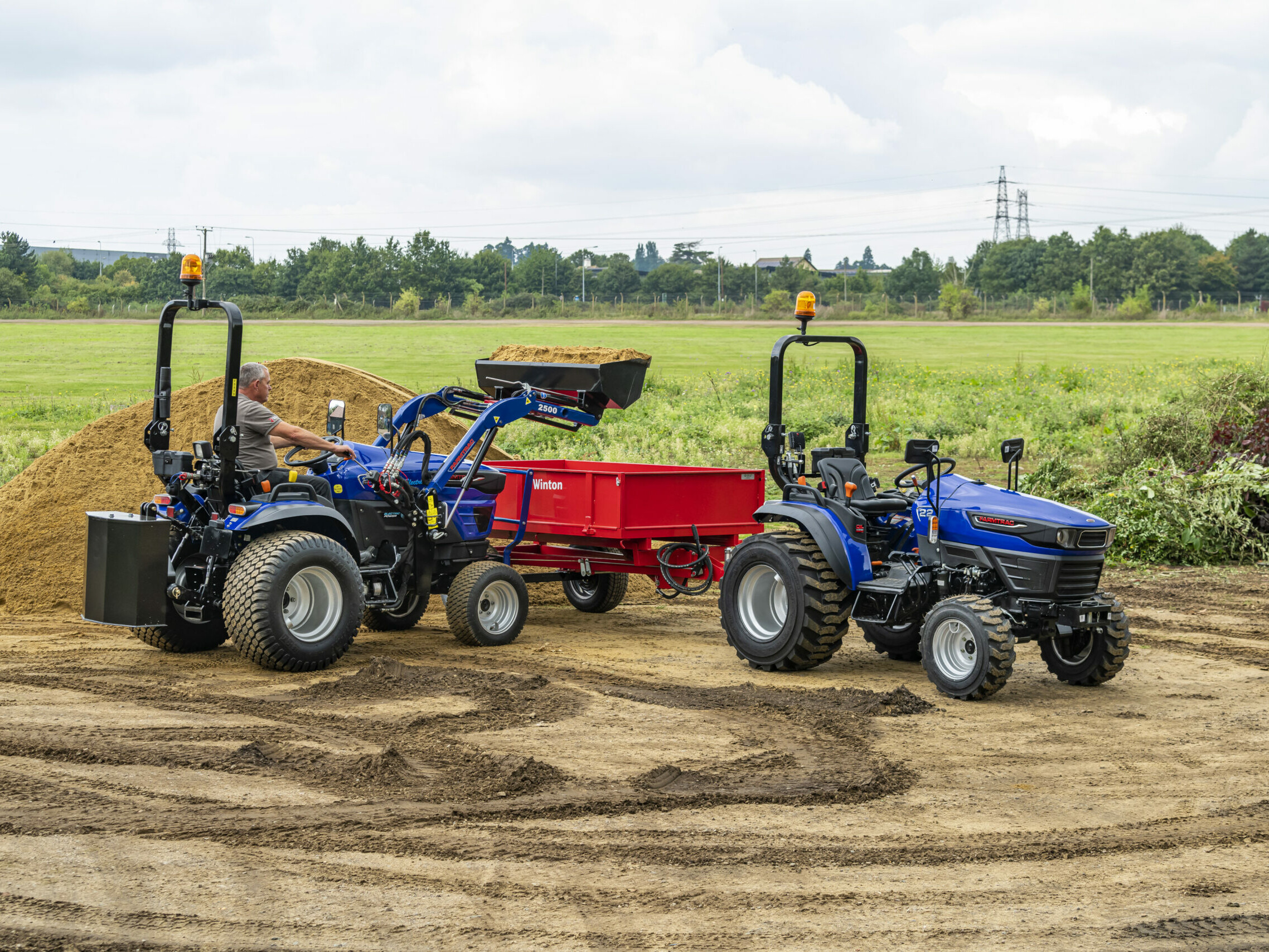 A Farmtrac FT25G being used with a front-loader to move sand, with a Farmtrac FT22 to the side.