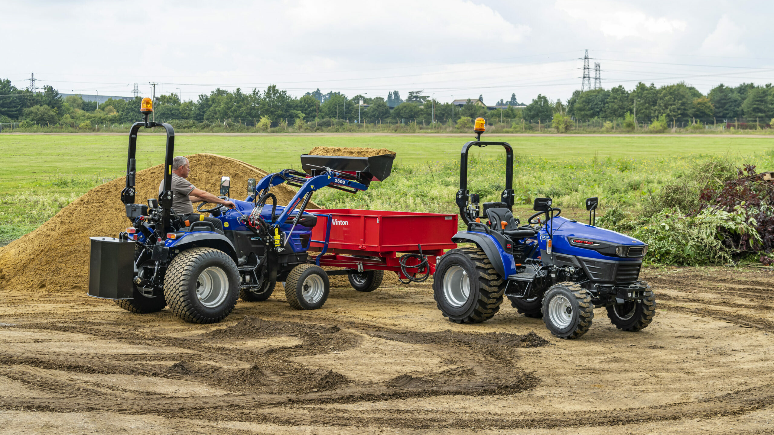 A Farmtrac FT25G being used with a front-loader to move sand, with a Farmtrac FT22 to the side.