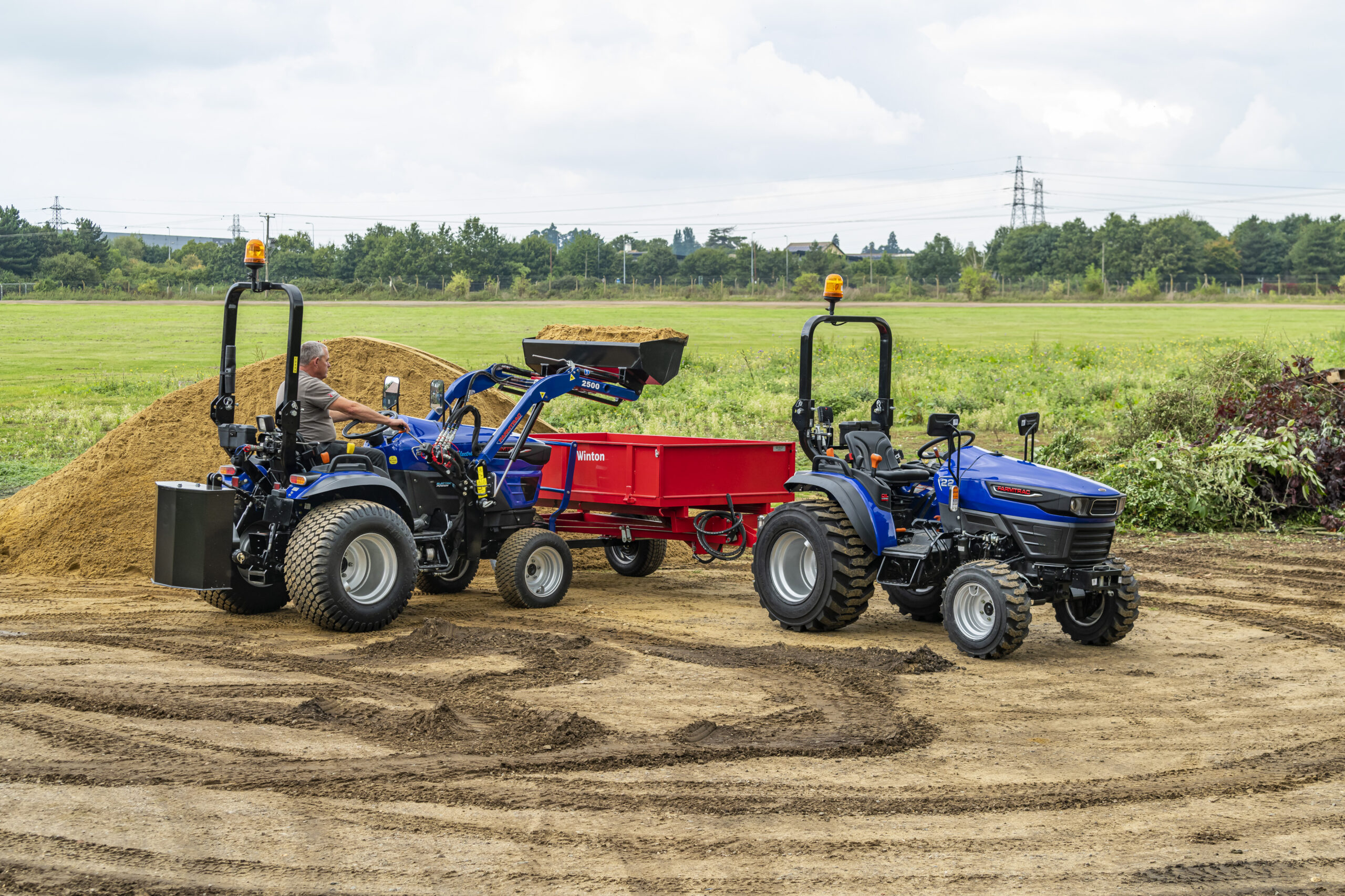 A Farmtrac FT25G being used with a front-loader to move sand, with a Farmtrac FT22 to the side.